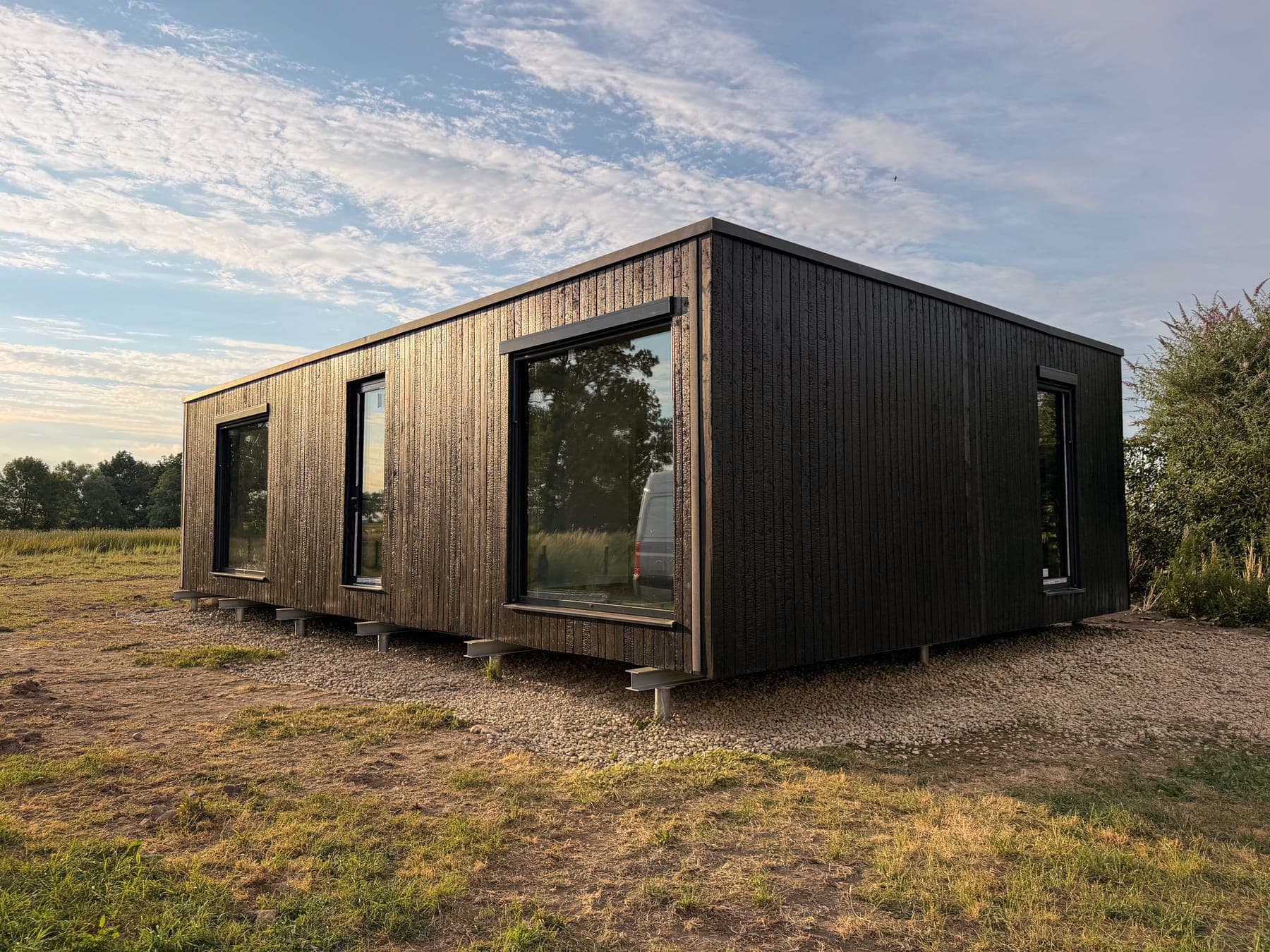 Serenity 95 m² on delivery day, in evening light. The edit removes delivery pallets, a protective tarp and the neighbouring yard with its fence and barn. The home, the light and the dry summer grass come from the camera original. (Edited with AI)