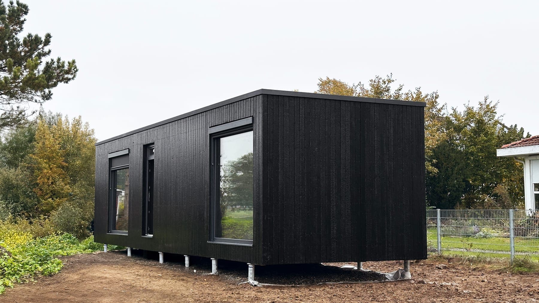 Wanderlust 48 m², photographed hours after delivery. The edit replaces mud, protective foil and a wire fence with finished gravel and greenery; the timber cladding, windows and ground screws come from the camera original. (Camera original)